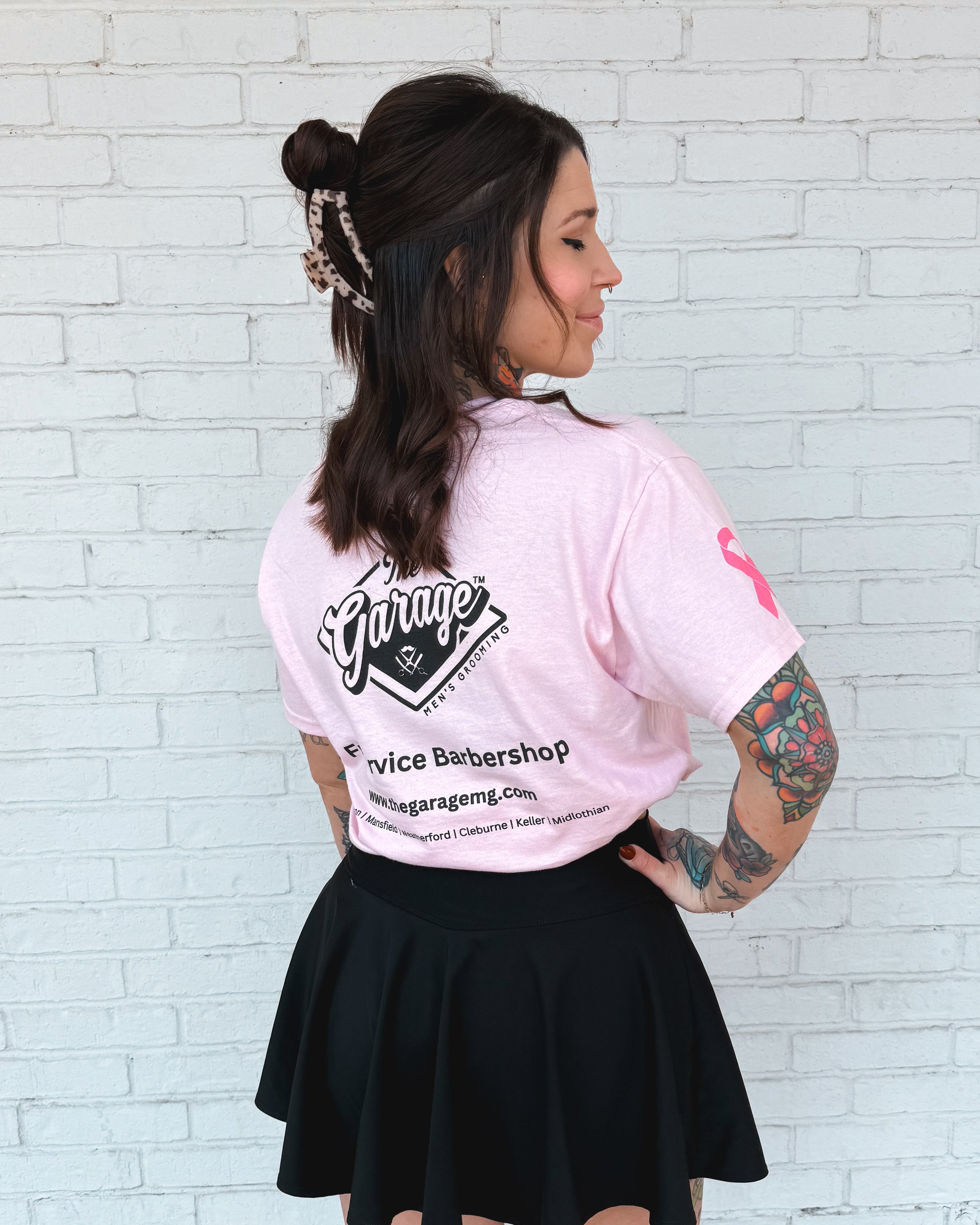Woman wearing a pink shirt with text and a black skirt against a white brick wall. Garage Men's Grooming Barbershop