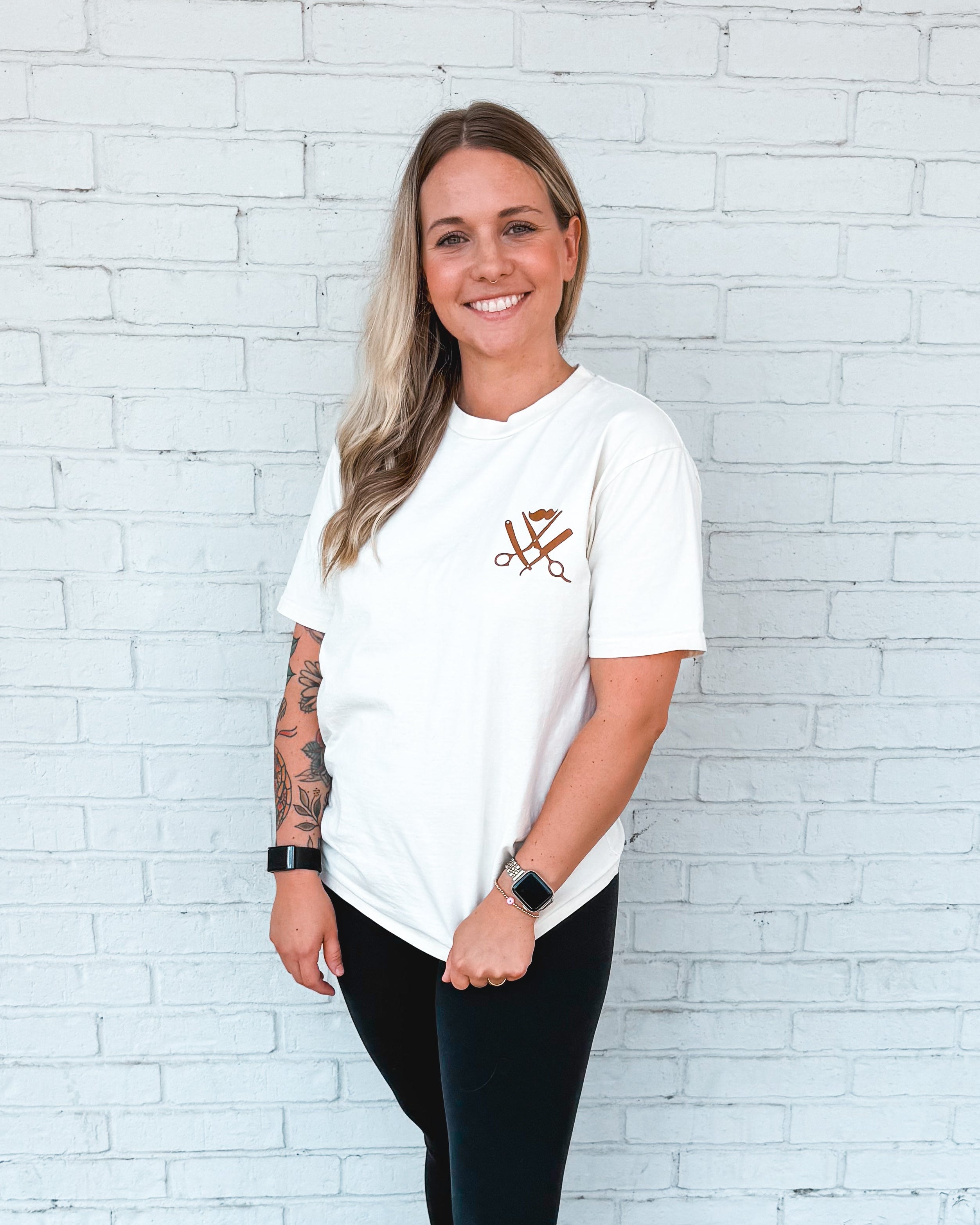 Woman wearing a white t-shirt with a logo and black leggings standing against a white brick wall. Garage Men's Grooming Merch
