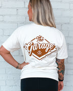 Person wearing a white t-shirt with a red 'The Garage' logo on the back against a white brick wall.