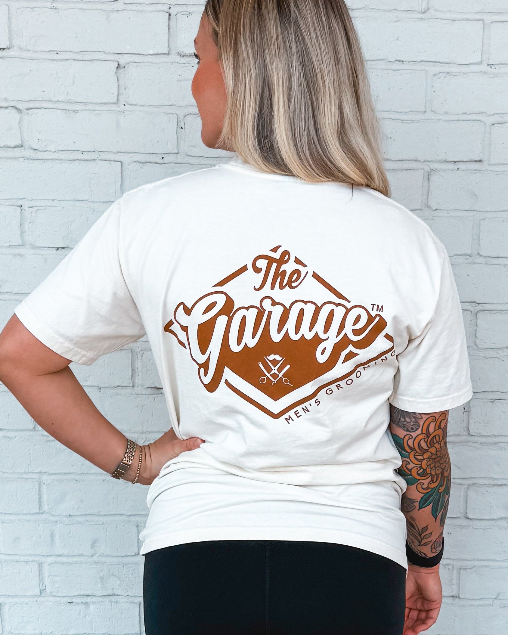 Person wearing a white t-shirt with a red 'The Garage' logo on the back against a white brick wall.